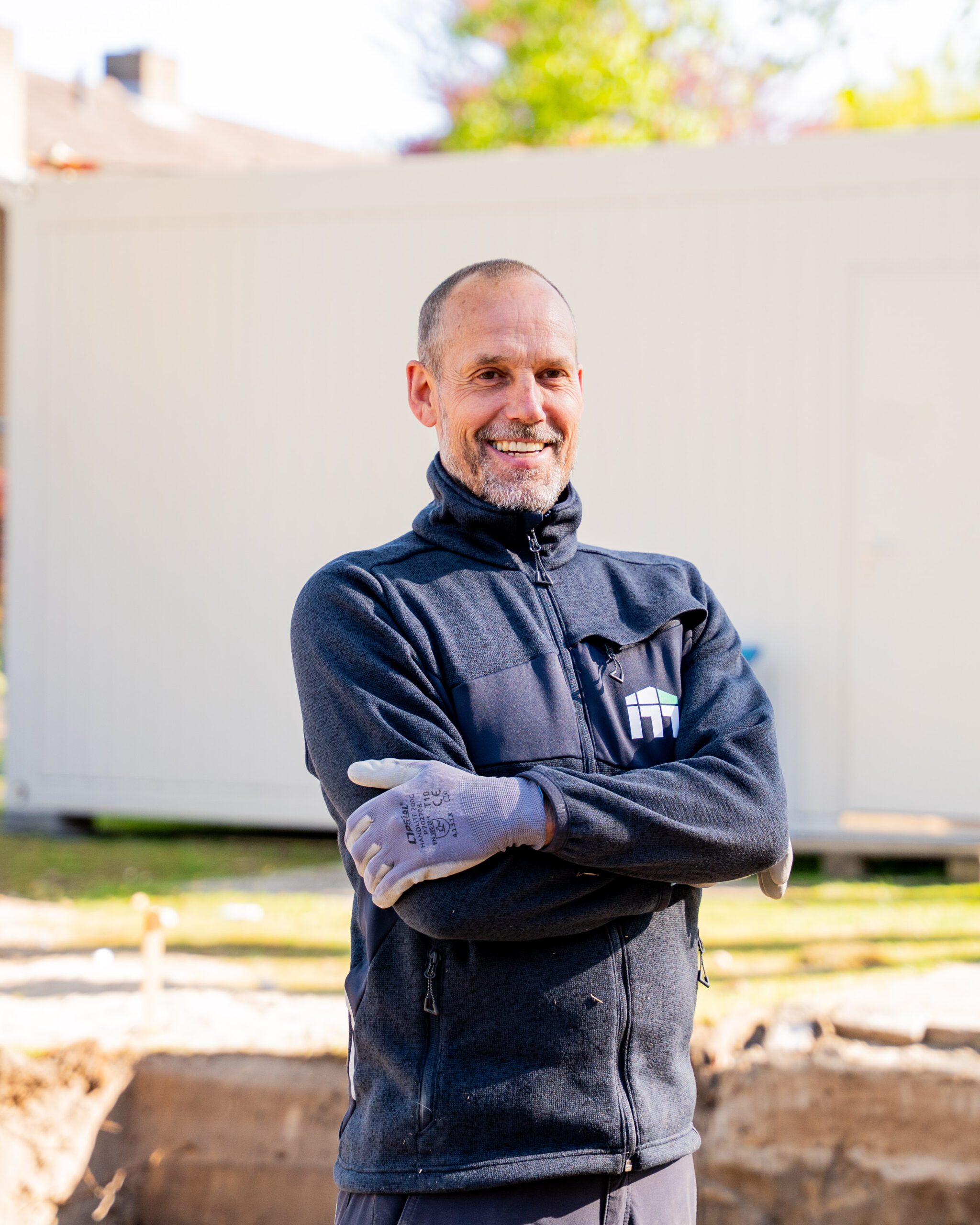 Smiling man in a zippered work jacket and gloves standing outdoors near a construction site.