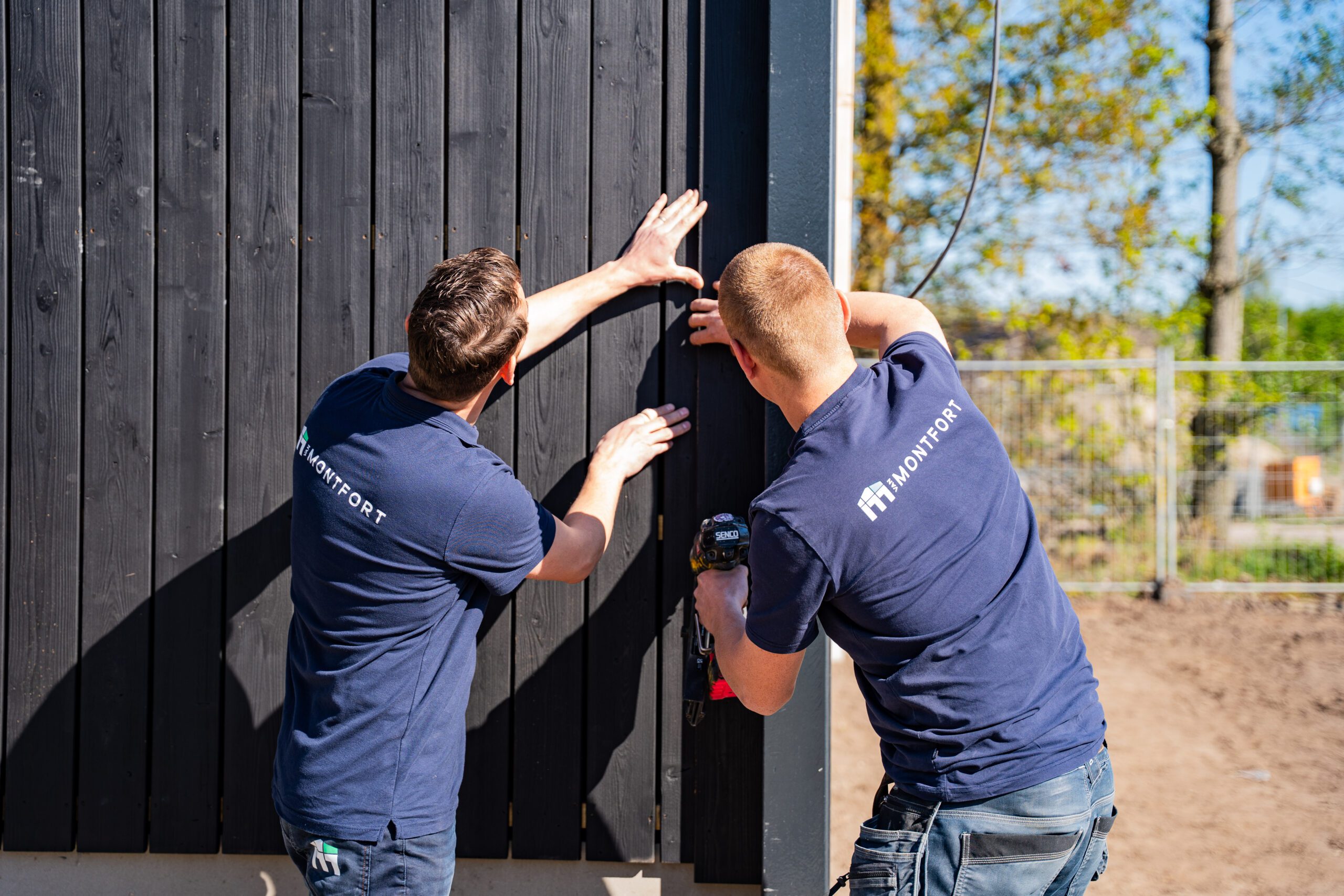 Two workers in navy shirts secure a tall dark wooden fence panel outdoors on a sunny day.