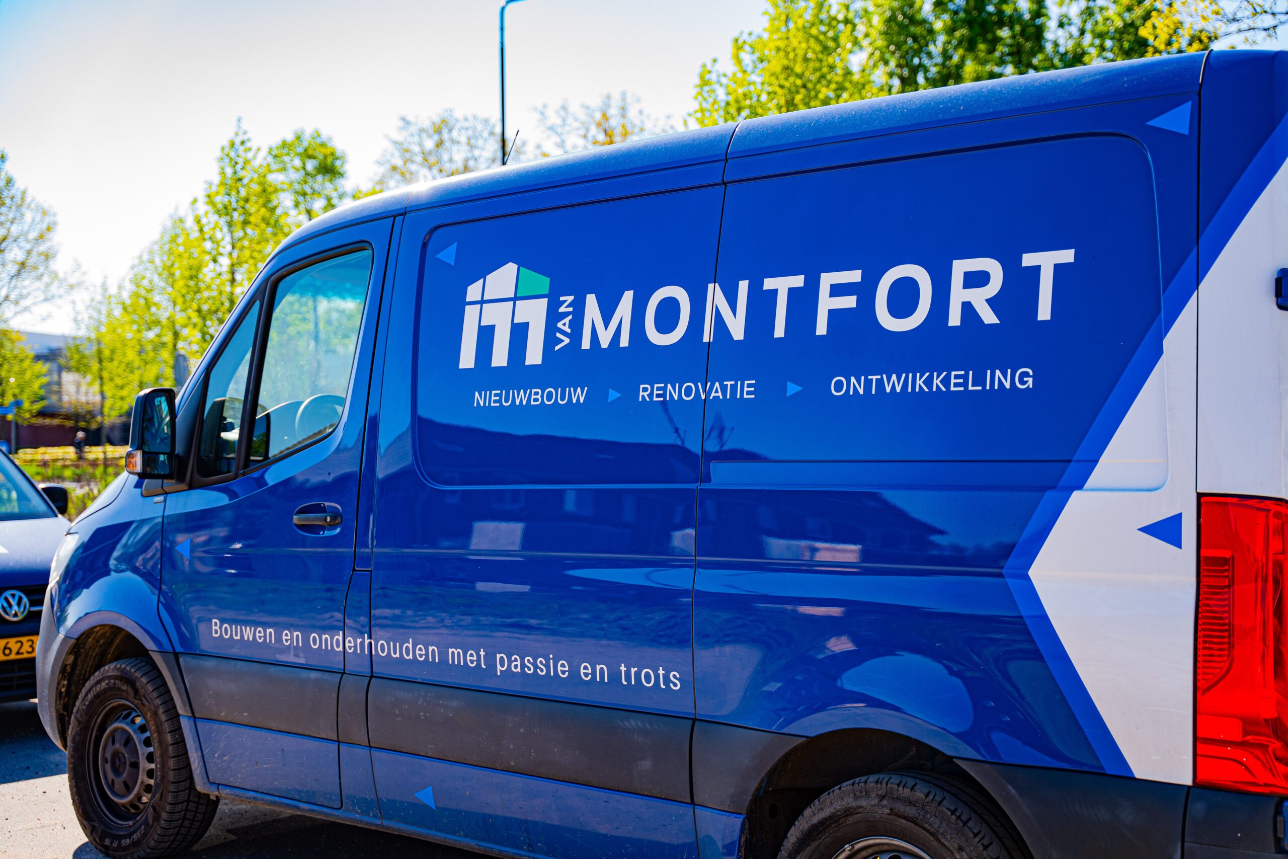 Blue company van with white text and logo on the side, showing 'MONFORT' and Dutch words, parked outdoors.