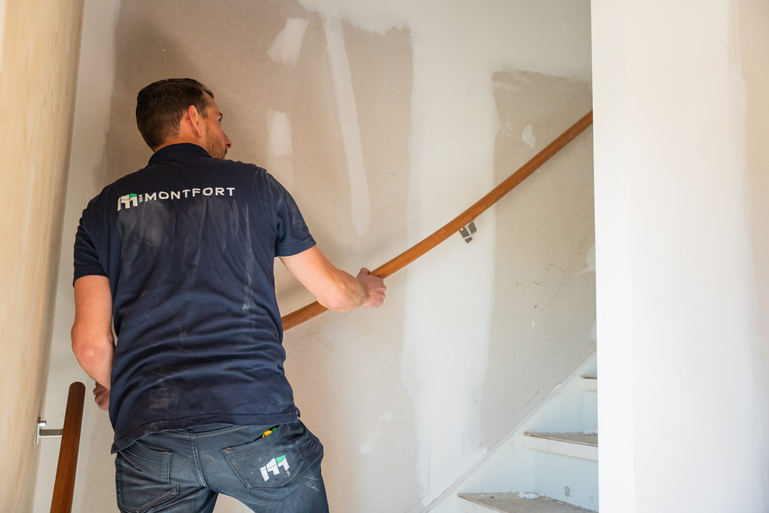 Man wearing a dark blue Montfort polo climbs a wooden staircase in a partially finished interior.