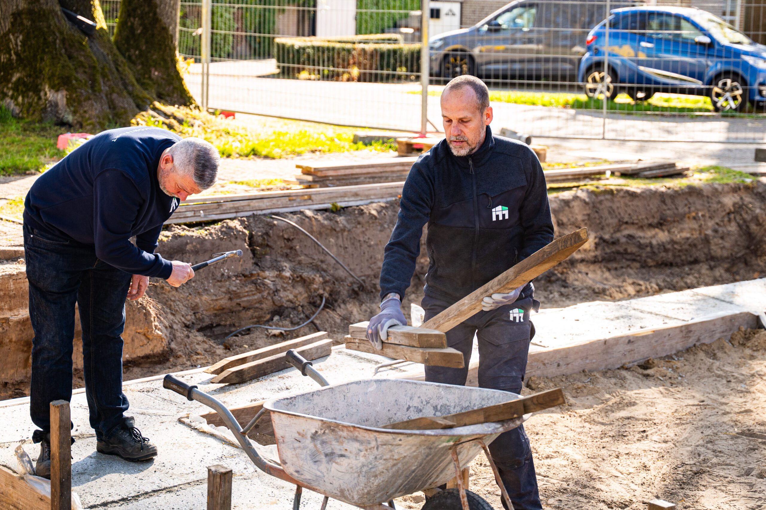 Two construction workers stand in a dug trench handling wooden planks near a wheelbarrow at an outdoor site