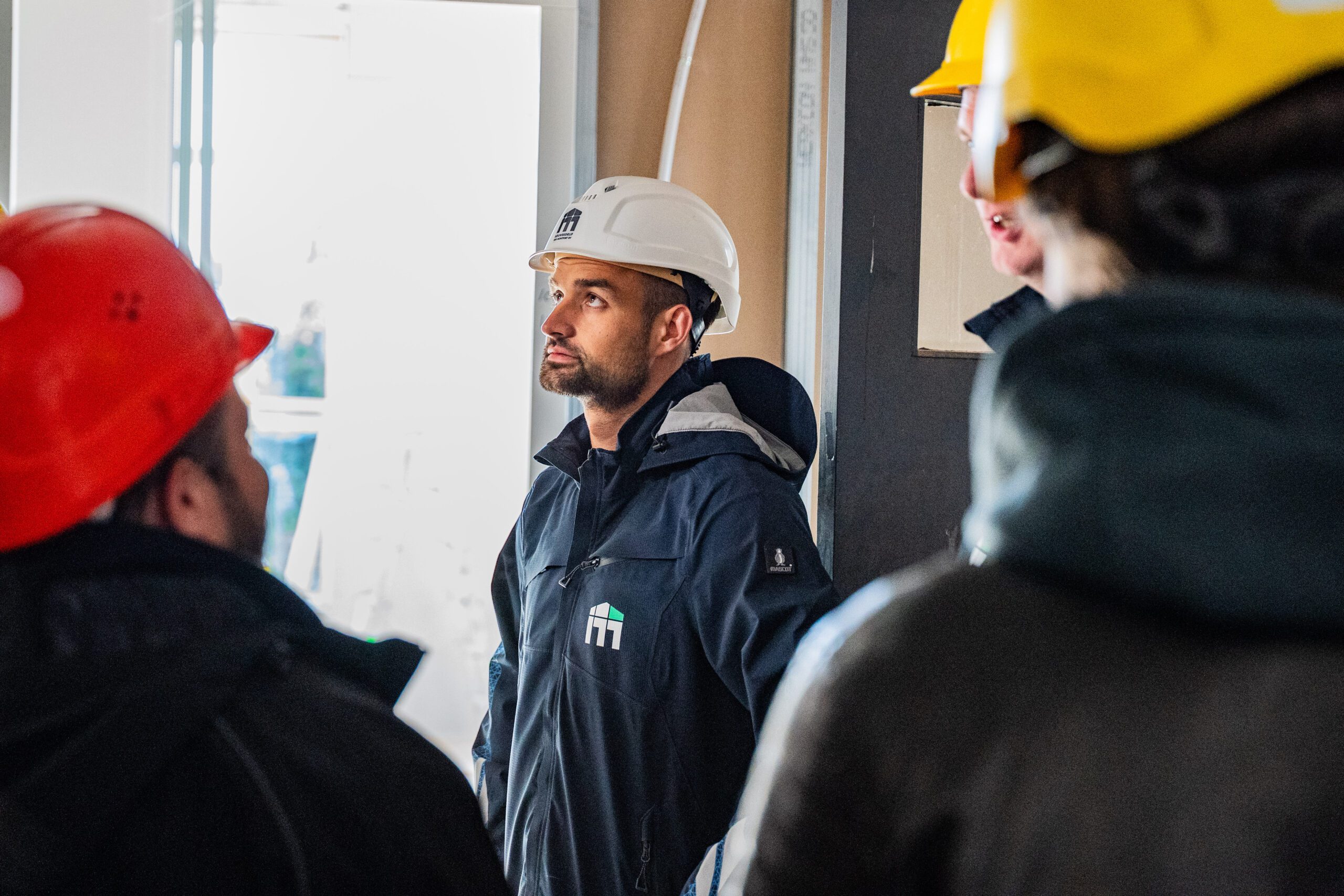 Construction worker in a white hard hat looks up while a group of teammates in red and yellow helmets listen nearby.