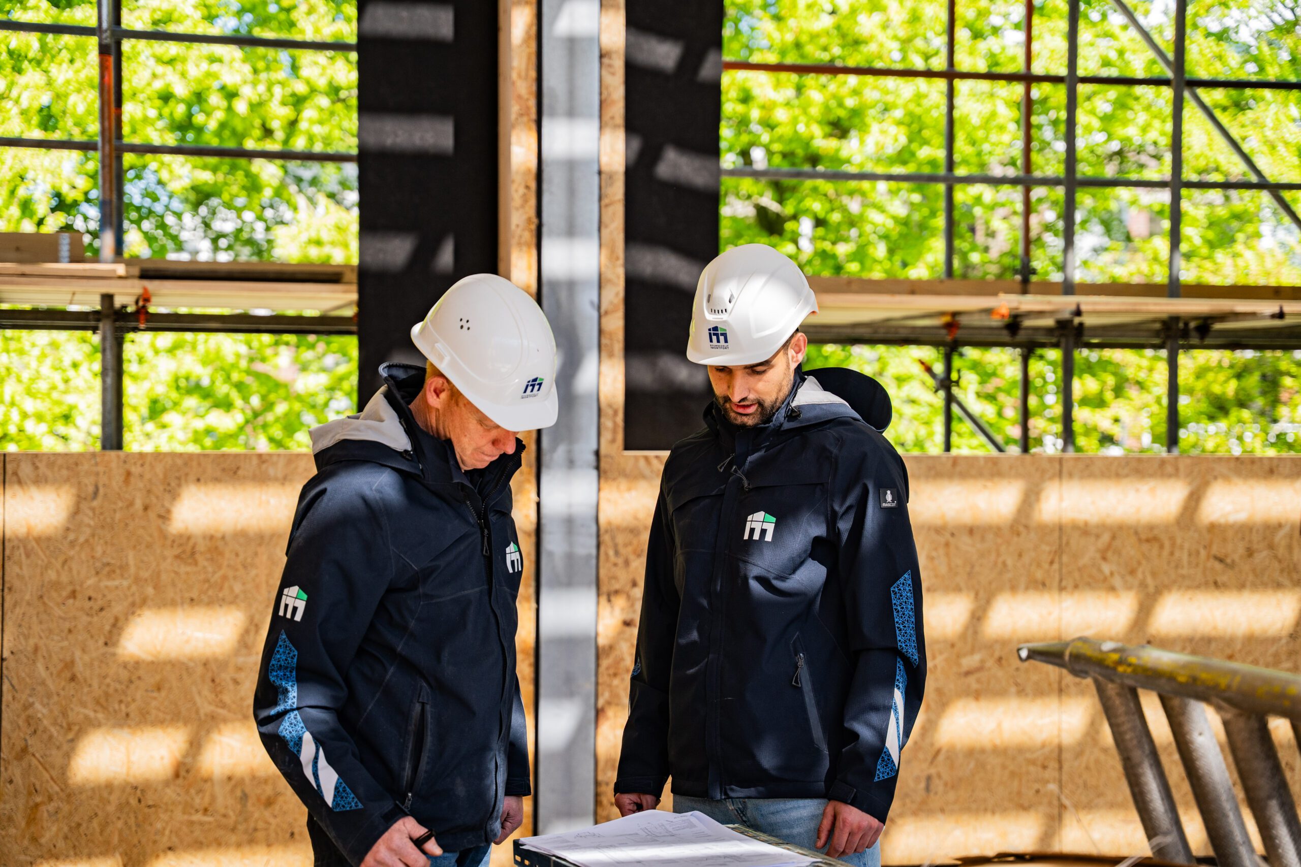Two construction workers in white hard hats study blueprints at an outdoor building site with wooden framing and green trees in the background.