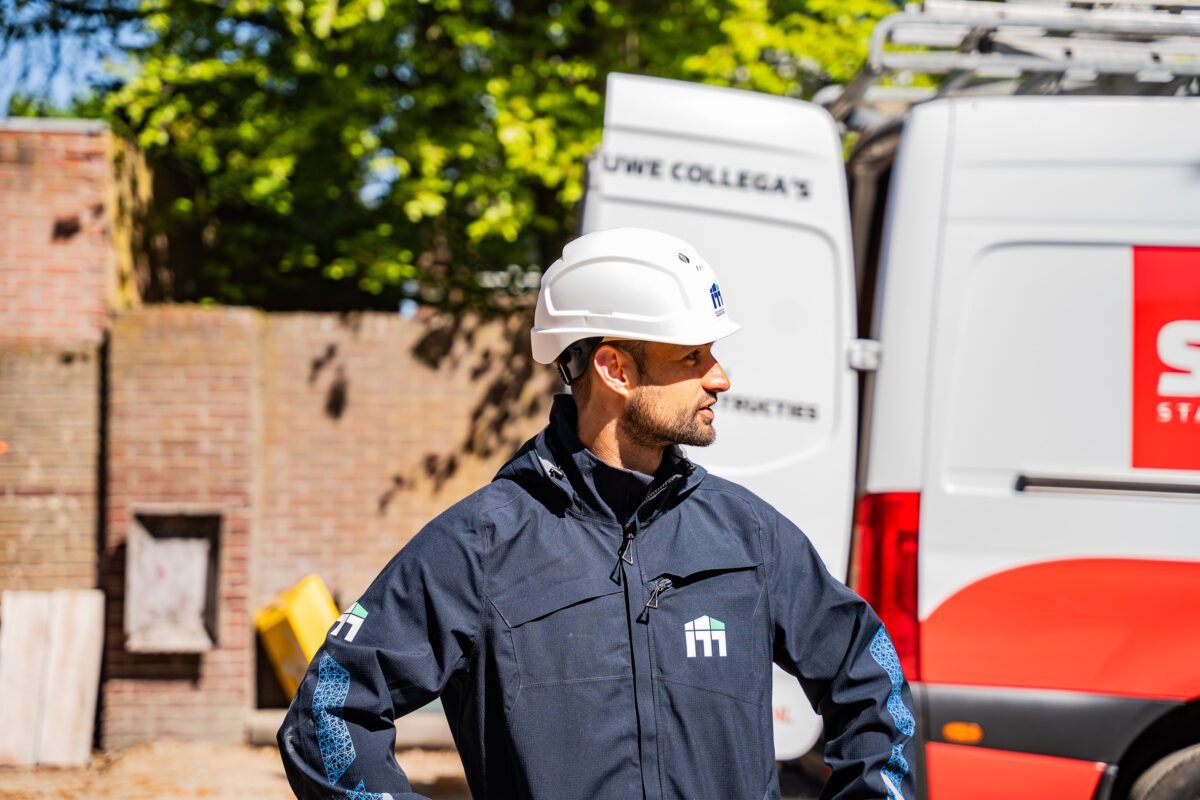 Construction worker in a white hard hat and dark jacket stands near a white van at a construction site with brick wall and greenery in the background.