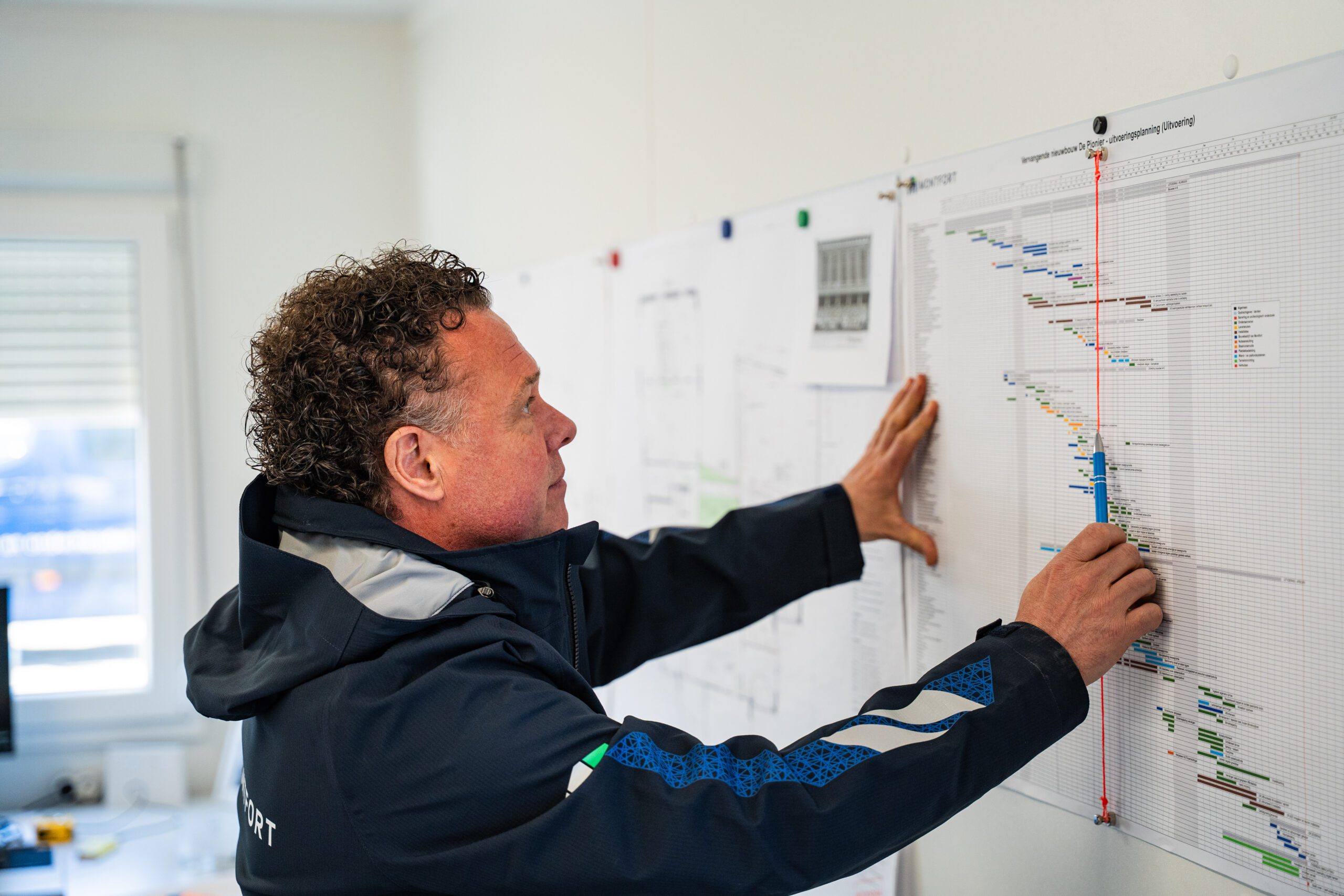 Man in a dark jacket studies a large wall-mounted project schedule with multicolored bars and a red vertical marker line, using his hands to point at it.