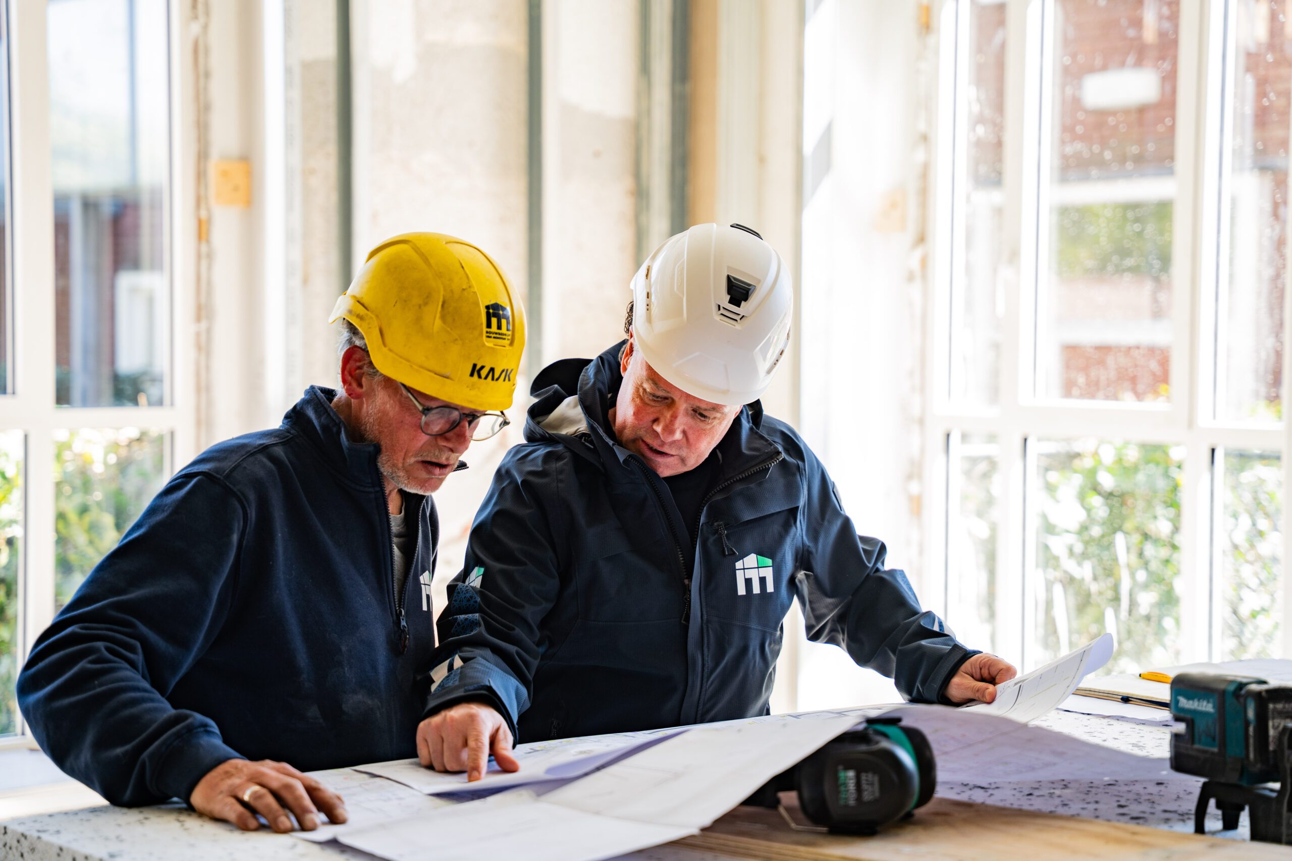 Two construction workers in helmets review blueprints at a bright construction site/office table.