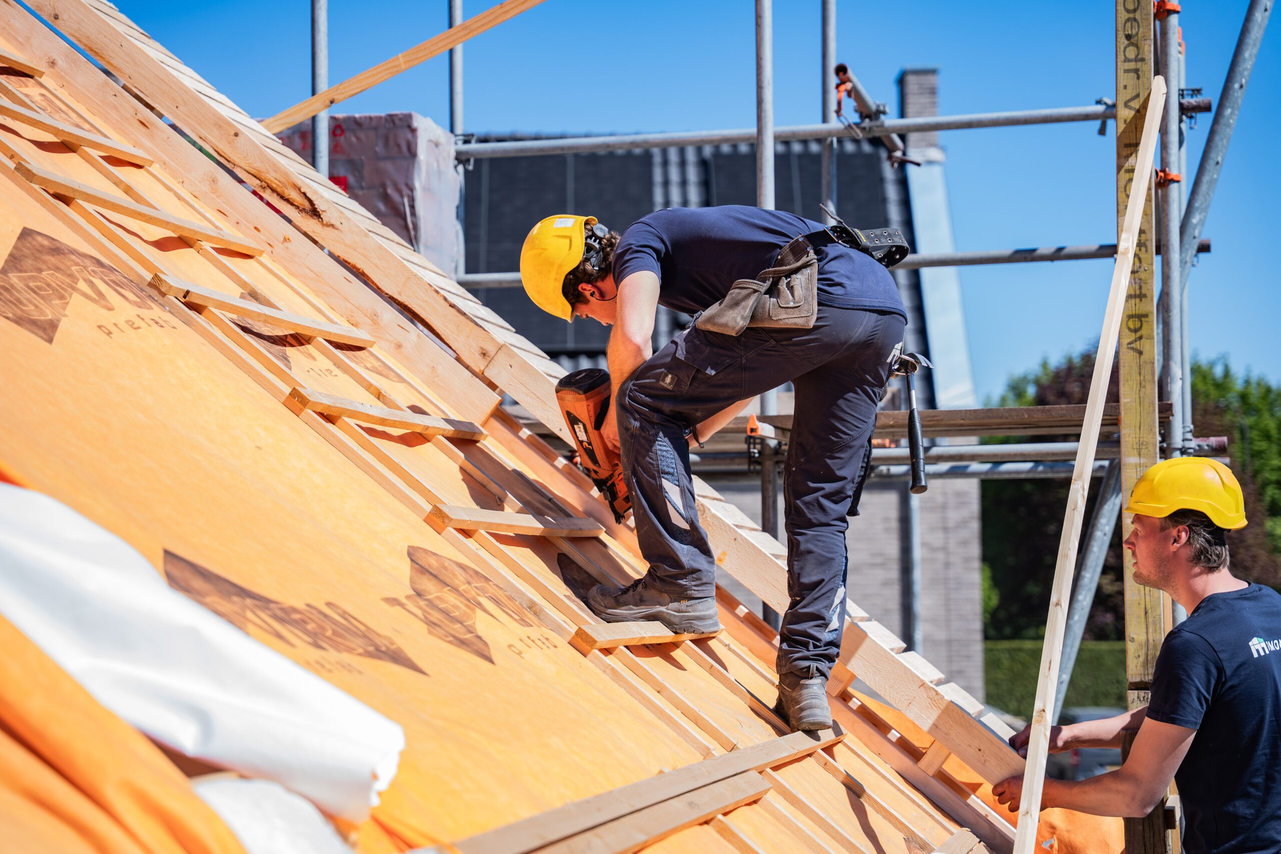 Construction workers install roof sheathing on a sunny job site, with wooden framing and scaffolding nearby, both wearing yellow hard hats.