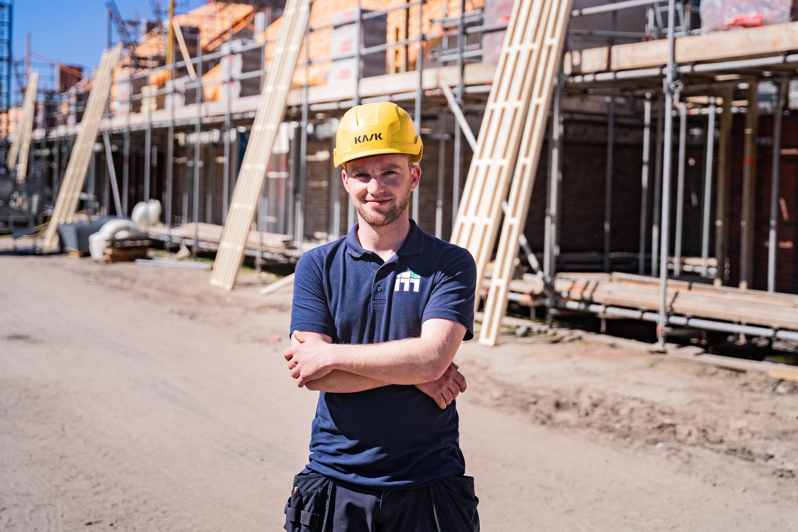 Construction worker in a blue polo with a yellow hard hat stands with arms crossed on a sunny building site.