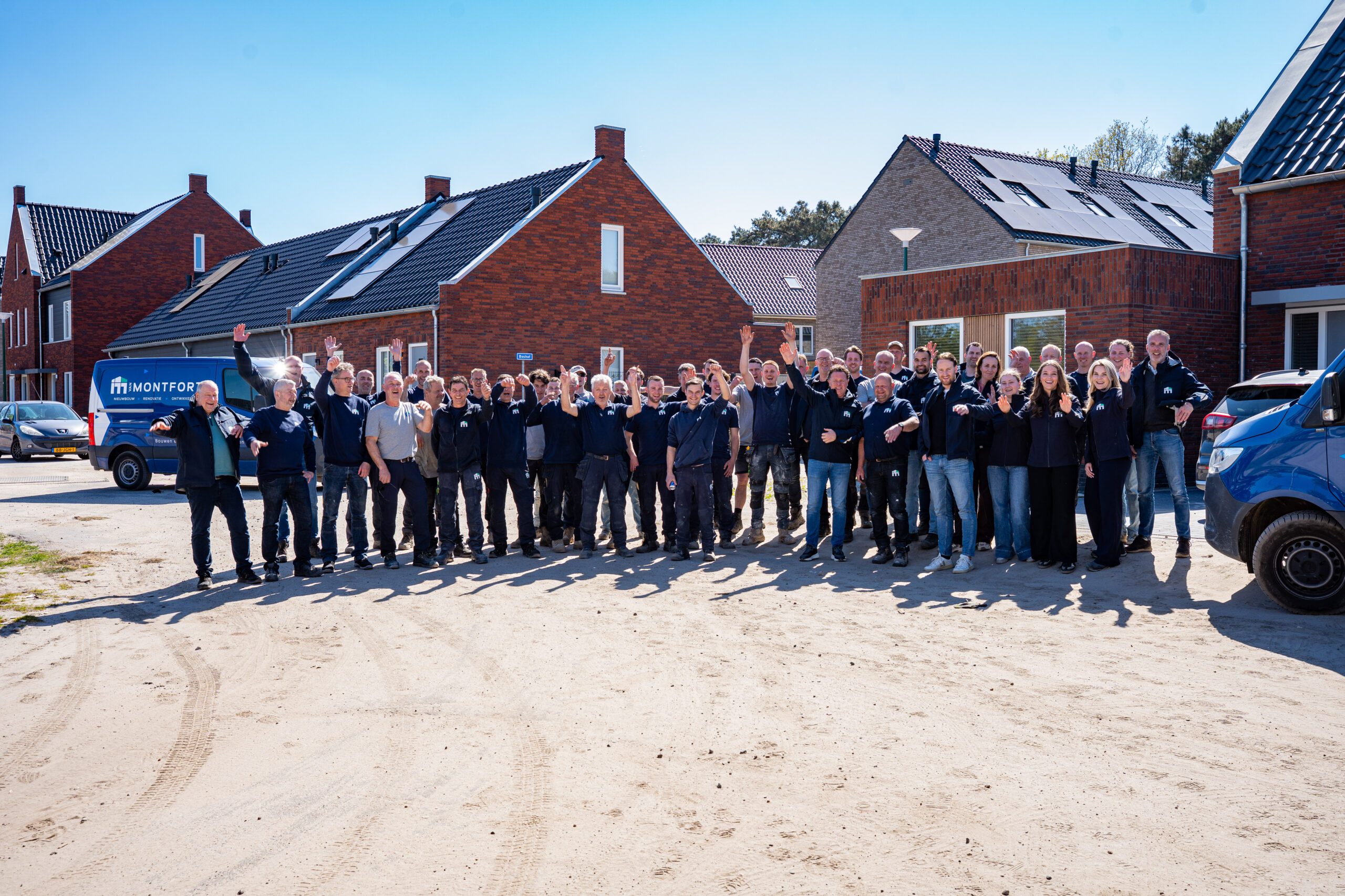 Large group of construction workers and staff waving hello in a sunny residential area with brick houses and company vans in the background